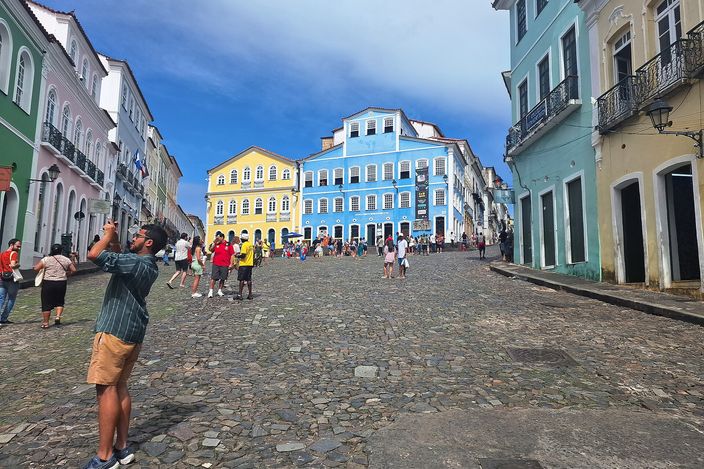 Largo do Pelourinho in Salvador seen from its top on a bright day: a broad cobbled square sloping downhill between rows of pastel colonial houses, the tall blue facade of the Fundacao Casa de Jorge Amado closing the view at the far end beside a yellow one, visitors scattered across the stones