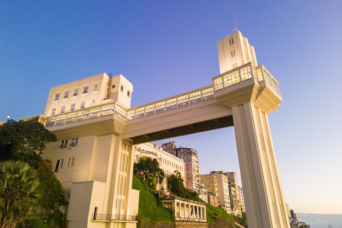 The Elevador Lacerda in Salvador photographed from below at golden hour: two tall cream art-deco towers joined near their tops by an enclosed walkway, the cliff of the upper city and its buildings behind them against a clear evening sky