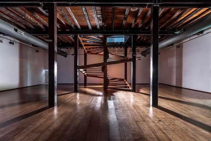 The interior hall of the Solar do Unhao in Salvador: a helical staircase of pale wooden treads rising through the centre of a dark timber-beamed hall, square black posts to either side and a worn plank floor catching the light from the windows