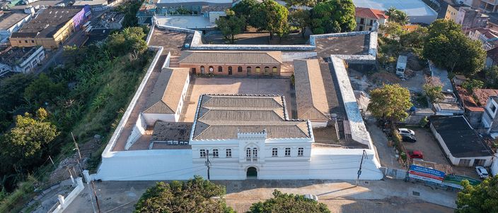 Aerial view in low evening light of the Forte de Santo Antonio Alem do Carmo in Salvador: a long whitewashed barracks range under terracotta roofs enclosing a walled rectangular courtyard, with the tiled roofs and trees of the Santo Antonio quarter pressing in on every side