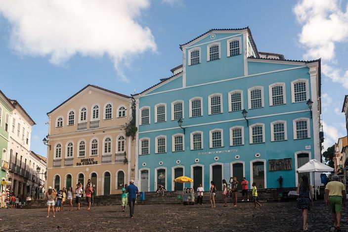 Sunlit pastel-and-blue colonial buildings with white shuttered windows rising above the cobbled slope of Largo do Pelourinho, people scattered across the square.