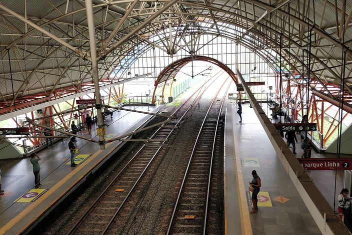 Elevated Salvador metrô platform under a curved steel-and-glass roof, twin tracks down the centre with passengers waiting and a yellow 'Linha 2' sign at right