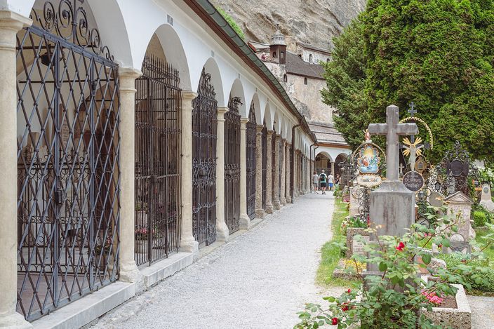 The arcaded family vaults of St Peter's cemetery in Salzburg: a row of wrought-iron grilles under white arches along the left, planted graves with a stone cross and flowers on the right, and the bare cliff face rising behind.