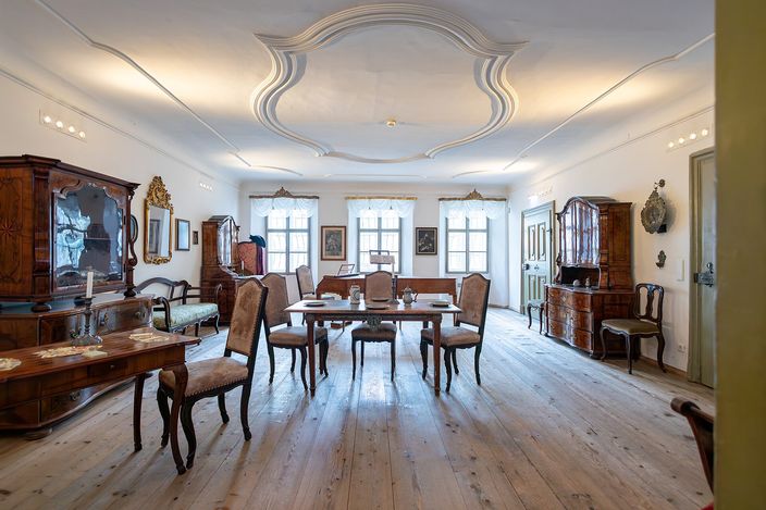 A restored eighteenth-century apartment inside Mozart's birthplace: a wide plank floor, a table with high-backed chairs under tall shuttered windows, walnut cabinets and framed pictures along the walls, and a moulded stucco medallion on the white ceiling.