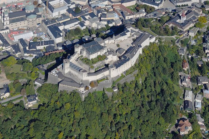 An aerial view of Hohensalzburg fortress along the spine of its wooded rock: the ring of ramparts and bastions enclosing courtyards and roofs around the tall white keep, with the roofs of Salzburg's old town at the top left.