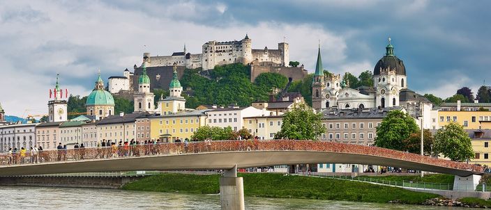 Salzburg's old town seen across the Salzach: the Marko-Feingold-Steg footbridge crossing the green river in the foreground, the pale baroque fronts, green domes and towers of the historic centre behind it, and Hohensalzburg fortress on its wooded rock above, under a grey sky.