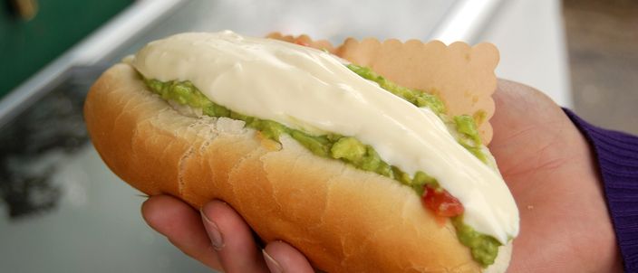 A hand holding a completo italiano: a soft hot-dog bun buried under mashed green avocado and a broad slick of white mayonnaise, with the sausage barely visible beneath.