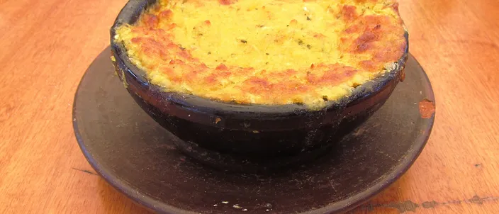 A pastel de choclo in a black fired-clay paila: the golden ground-sweetcorn topping baked until browned on top, served on a plate on a wooden table.