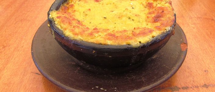A pastel de choclo in a black fired-clay paila: the golden ground-sweetcorn topping baked until browned on top, served on a plate on a wooden table.