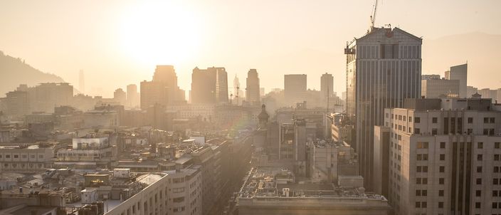 Golden backlit view over downtown Santiago's mid-rise blocks and glass towers, the Andes foothills fading into haze under a hazy sun