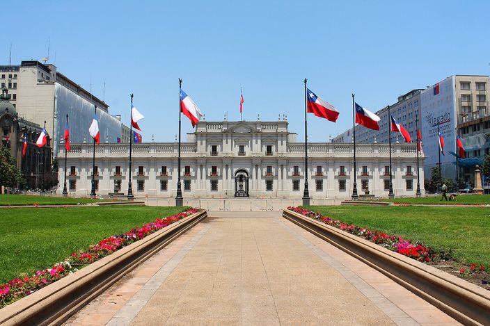 Palacio de La Moneda seen head-on across a paved plaza lined with red and white flowerbeds, a double row of Chilean flags on tall poles leading the eye to the long neoclassical white facade under a clear blue sky