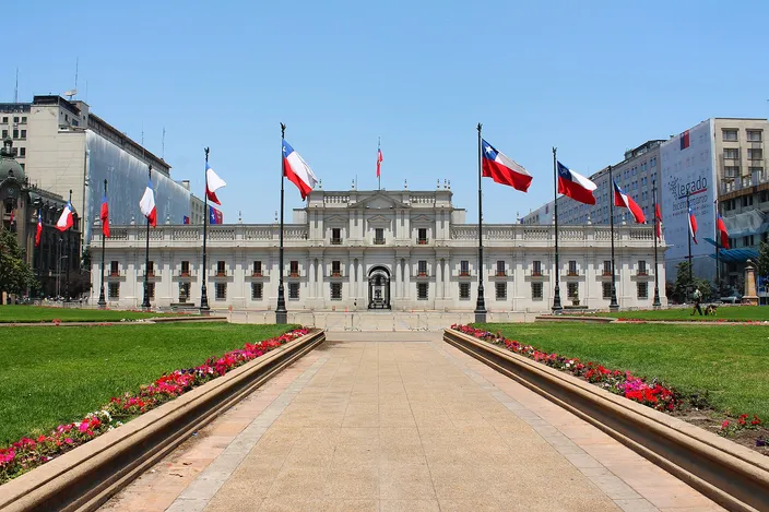 Palacio de La Moneda seen head-on across a paved plaza lined with red and white flowerbeds, a double row of Chilean flags on tall poles leading the eye to the long neoclassical white facade under a clear blue sky