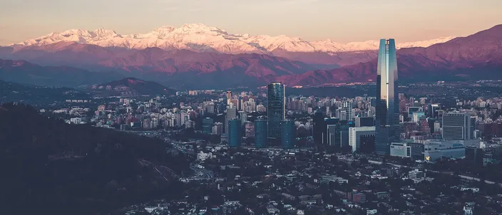 Santiago at dusk seen from a dark wooded hillside: a dense field of low-rise blocks with a cluster of glass towers and one tall pale skyscraper rising above them, backed by a long wall of snow-capped Andes under a pink and gray sky