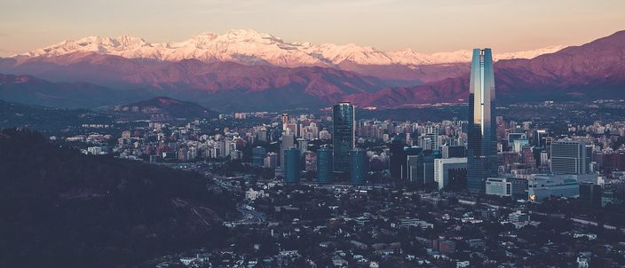 Santiago at dusk seen from a dark wooded hillside: a dense field of low-rise blocks with a cluster of glass towers and one tall pale skyscraper rising above them, backed by a long wall of snow-capped Andes under a pink and grey sky