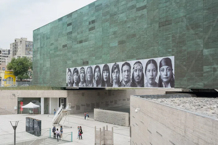 The Museo de la Memoria y los Derechos Humanos: a large windowless block clad in oxidised green copper, a long horizontal band of black-and-white portrait photographs mounted across its face, and the raised plaza and steps below with a few people crossing