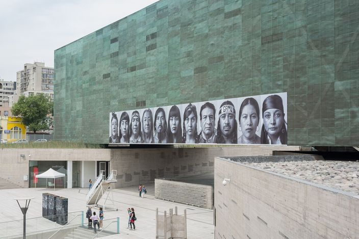 The Museo de la Memoria y los Derechos Humanos: a large windowless block clad in oxidised green copper, a long horizontal band of black-and-white portrait photographs mounted across its face, and the raised plaza and steps below with a few people crossing