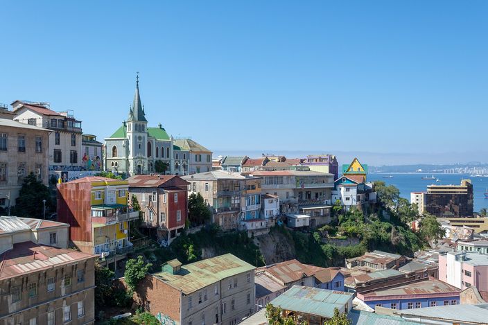 Cerro Concepción in Valparaíso seen from the neighbouring hill: painted timber and metal-clad houses stacked along a cliff edge, a green Gothic church spire rising among them, and the bay with anchored ships beyond