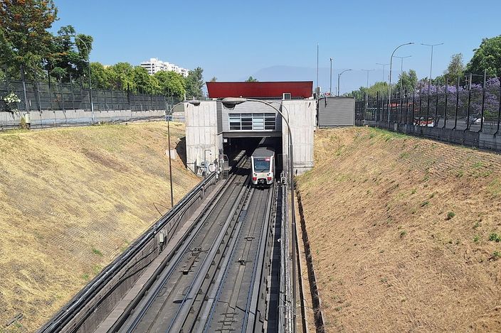A Santiago Metro Line 1 train emerging from a tunnel portal into an open cutting, twin tracks running toward the camera, dry grassy banks either side and the Andes faintly visible on the horizon under a clear sky