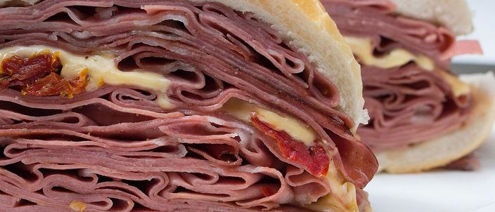 A mortadella sandwich cut in half on a white plate, showing a thick pile of folded pink mortadella slices with melted cheese inside a crusty bread roll.