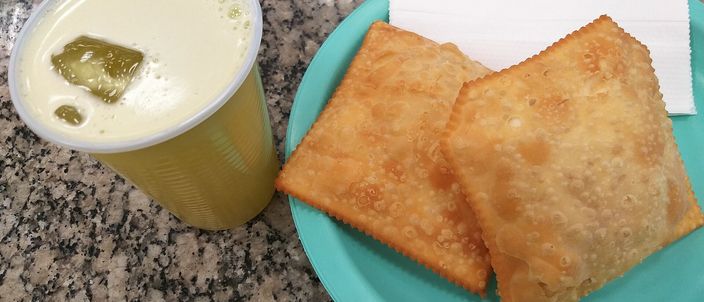 Golden, blistered rectangular pastéis on a plate beside a cup of pale sugarcane juice, on a granite market counter.