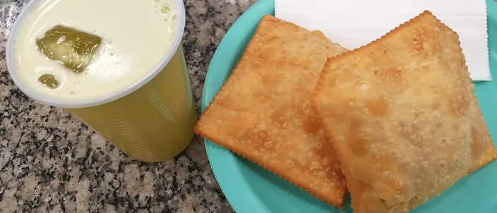 Golden, blistered rectangular pastéis on a plate beside a cup of pale sugarcane juice, on a granite market counter.