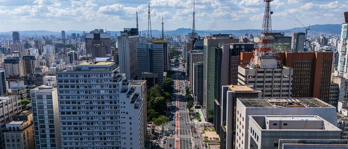 Aerial view down Avenida Paulista, São Paulo — a broad avenue lined with dense high-rise towers stretching to the horizon, twin TV masts on the ridge, under a bright blue sky with scattered cloud