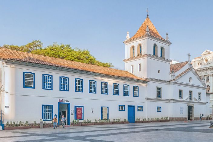 A low whitewashed colonial building with blue shutters and a red-tiled roof beside a white church with a square bell tower topped by a small dome, an empty paved square in front under a clear blue sky.