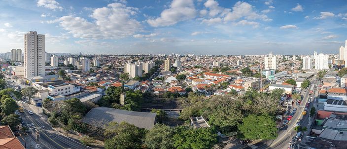A city stretching to the horizon under tall white clouds: apartment towers on the left, a spread of low roofs and green trees in the middle, a broad avenue with traffic along the bottom right.