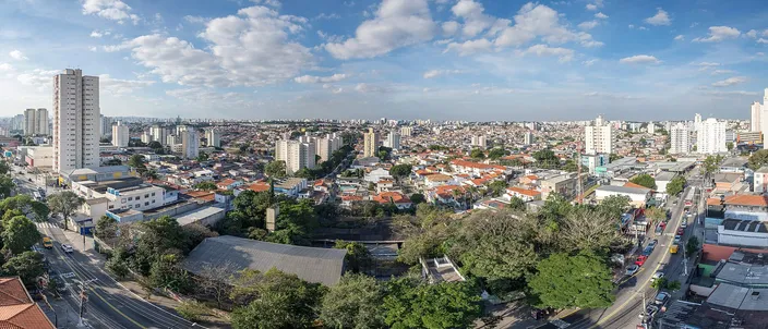 A city stretching to the horizon under tall white clouds: apartment towers on the left, a spread of low roofs and green trees in the middle, a broad avenue with traffic along the bottom right.