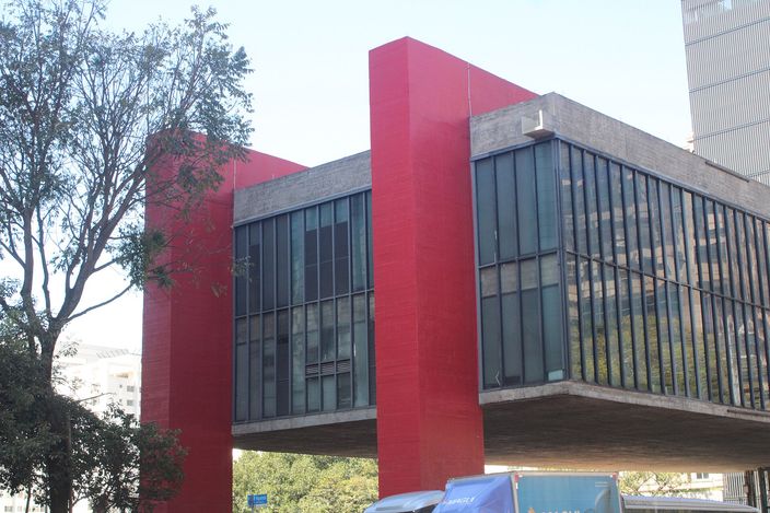 The Museu de Arte de Sao Paulo from the pavement: a long glass-and-steel box suspended between two pairs of bright red concrete piers, the open space beneath it empty, trees to the left.