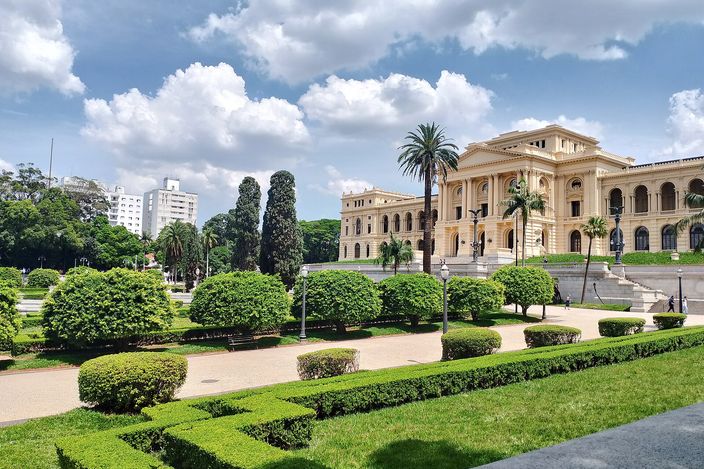 The Museu do Ipiranga at the head of its formal garden: a long cream-and-yellow nineteenth-century palace with a central pediment, seen across clipped box hedges, round shrubs and palms under a blue sky with white cloud.