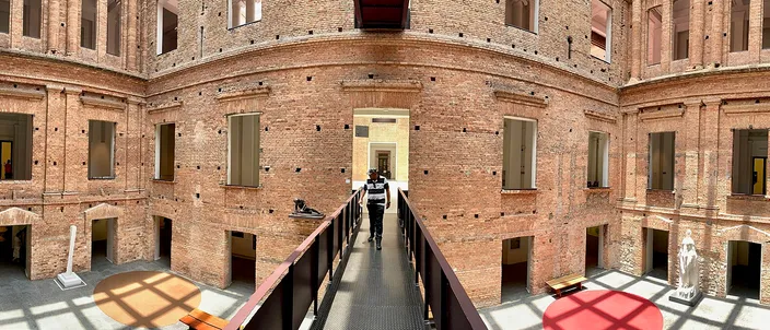 A sunlit brick courtyard inside the Pinacoteca: unrendered nineteenth-century brick walls with shuttered windows on all sides, a steel walkway crossing the space at upper-floor level with a single figure walking along it, under a glass roof.