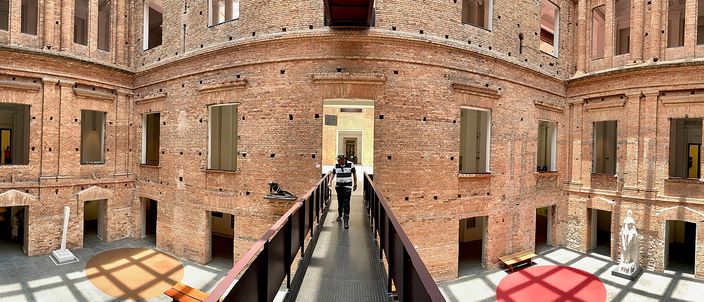 A sunlit brick courtyard inside the Pinacoteca: unrendered nineteenth-century brick walls with shuttered windows on all sides, a steel walkway crossing the space at upper-floor level with a single figure walking along it, under a glass roof.