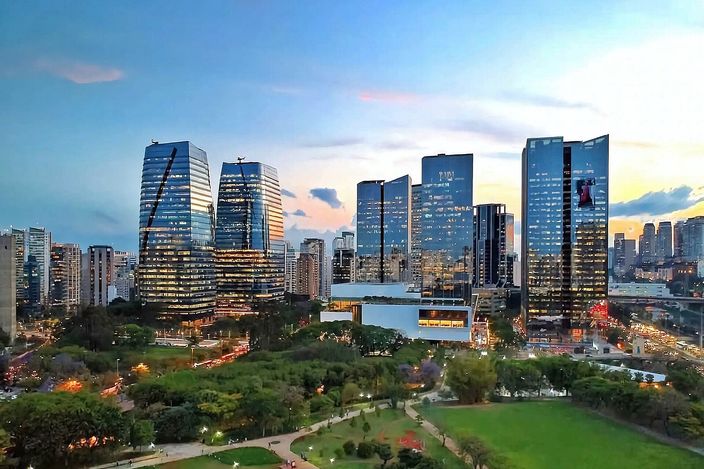 Cluster of curved glass office towers glowing at sunset above a large green park, with a wider low-rise city skyline stretching to the horizon