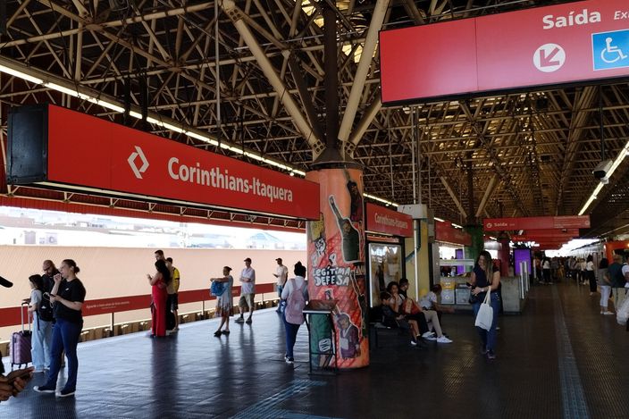Busy elevated CPTM/Metro platform at Corinthians-Itaquera, red 'Corinthians-Itaquera' and 'Saída' signage under a steel-truss roof, passengers waiting and walking along the concourse with a train at the platform.