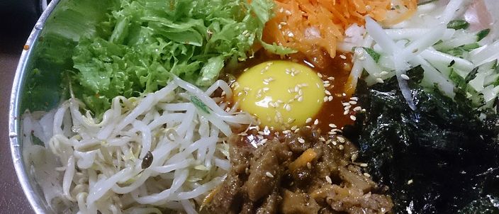 A bowl of bibimbap before mixing: white rice topped with neatly arranged separate mounds of seasoned vegetables — carrot, white radish, bean sprouts, lettuce, seaweed — around a central raw egg yolk