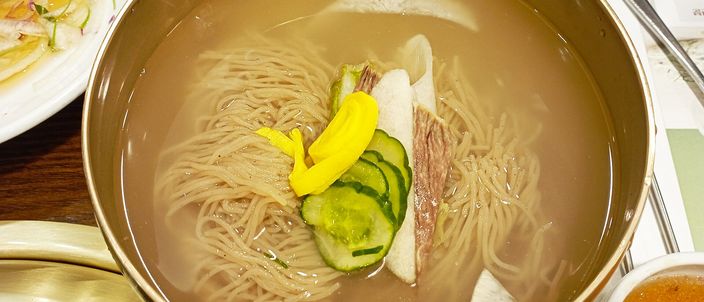 A brass bowl of mul naengmyeon: thin grey buckwheat noodles in a clear cold broth, topped with sliced cucumber, yellow pickled radish and slices of beef