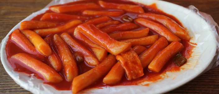 A plate of tteokbokki: cylindrical white rice cakes coated in a glossy red chili sauce, with slices of fish cake