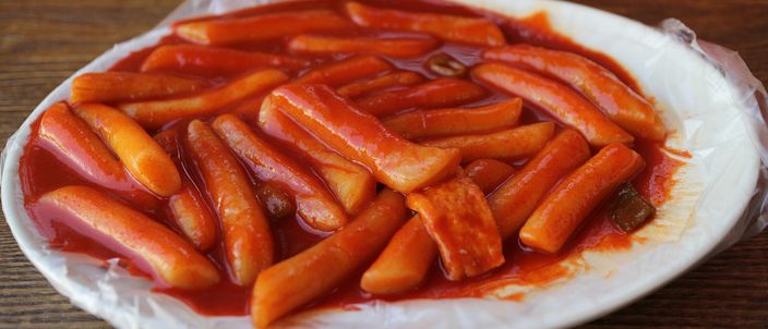 A plate of tteokbokki: cylindrical white rice cakes coated in a glossy red chilli sauce, with slices of fish cake