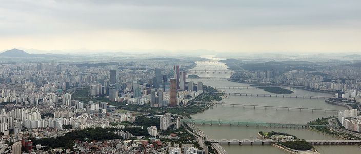 Aerial view of Seoul with the Han River curving through a dense sea of high-rises, multiple bridges spanning the water and mountains fading into haze on the horizon