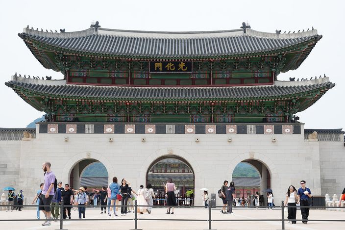 The double-roofed wooden Gwanghwamun gate seen head-on across a broad paved terrace, its three stone arches open and small groups of people walking in front of it