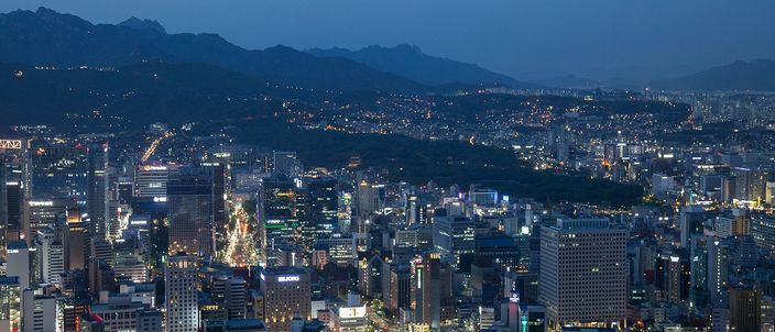 A dense city centre at blue hour seen from high ground, office towers and lit streets filling the basin below and a ridge of rocky mountains fading into the dusk behind it