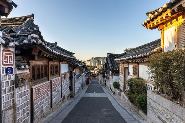 A narrow paved lane running downhill between rows of single-storey tiled traditional Korean houses with white walls and patterned brickwork, empty of people, with modern tower blocks visible far below at the end of the street