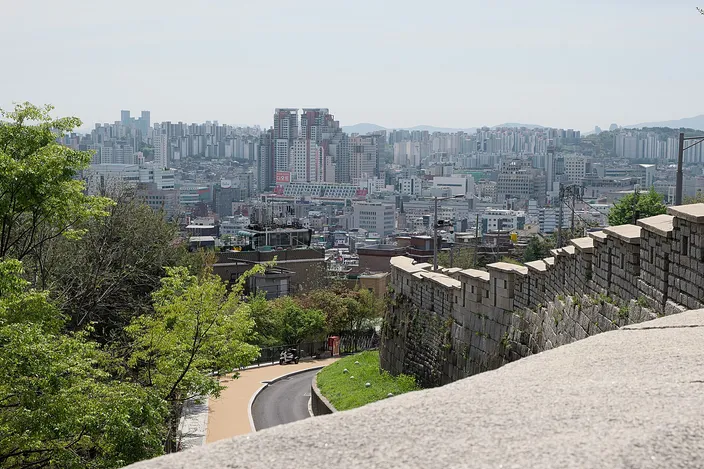 A high stone city wall with a crenellated top running along a ridge, a footpath curving below it, and a dense low-rise city of apartment blocks and small buildings stretching to hills on the horizon