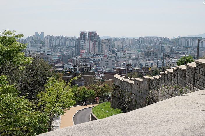 A high stone city wall with a crenellated top running along a ridge, a footpath curving below it, and a dense low-rise city of apartment blocks and small buildings stretching to hills on the horizon
