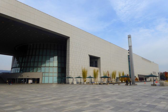 A very long, low stone-clad modern museum building with a glazed recessed entrance at one end, seen across a wide empty paved forecourt with small trees and parasols along its base