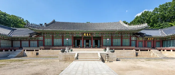 A long single-story Korean palace hall with a deep tiled double roof and green-and-red painted eaves, seen head-on and symmetrically across an empty stone forecourt under a clear blue sky