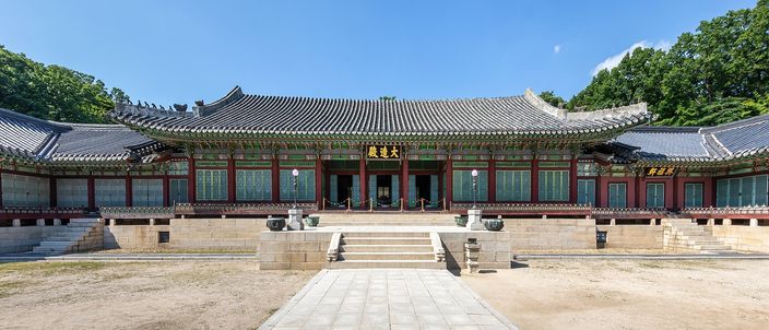 A long single-storey Korean palace hall with a deep tiled double roof and green-and-red painted eaves, seen head-on and symmetrically across an empty stone forecourt under a clear blue sky