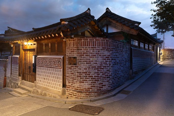 A traditional Korean hanok house on a sloping corner at blue hour in Bukchon, with a tiled curved roof, wooden gate, patterned brick wall and a quiet empty lane