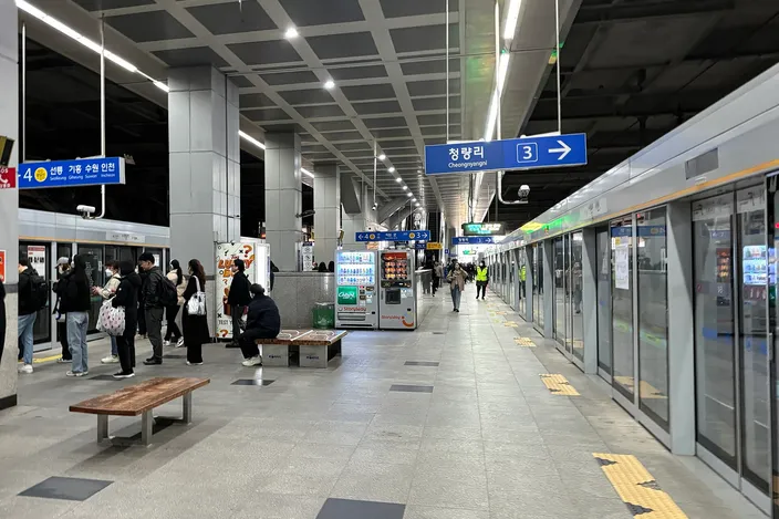 A Seoul subway platform with people seated and standing on a bench, platform screen doors, a blue directional sign reading Cheongnyangni, vending machines, and a train stopped alongside.